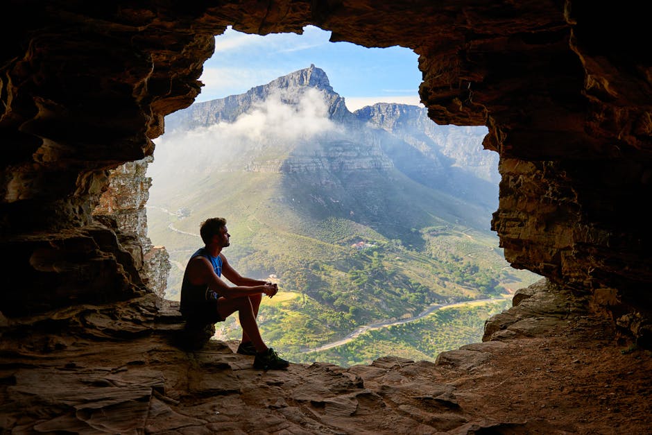 A man sitting in a cave overlooking a majestic mountain landscape under daylight